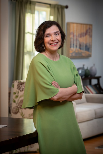 Person wearing a green dress, standing with arms crossed near a table in a softly lit living room; a window with curtains, a painting, and home decor appear in the background